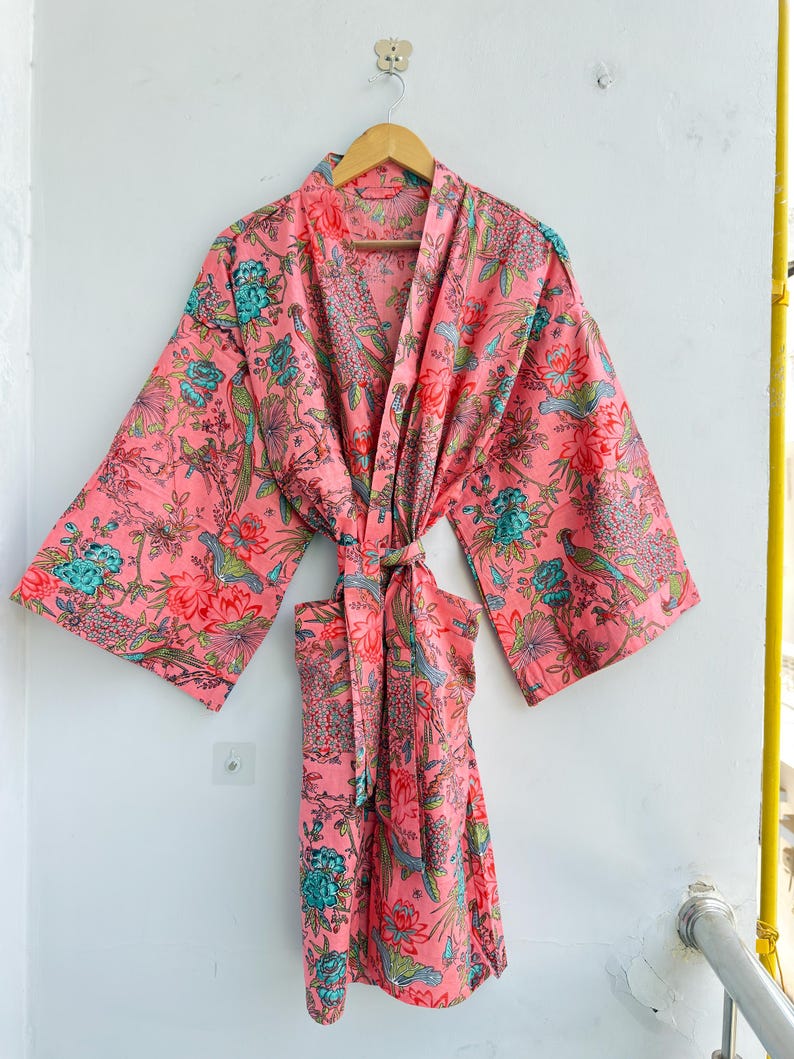 May include: A long-sleeved kimono robe in a vibrant coral pink base colour, featuring an intricate floral and bird pattern in shades of teal, green, and red. The garment includes a matching fabric tie belt at the waist and wide sleeves.