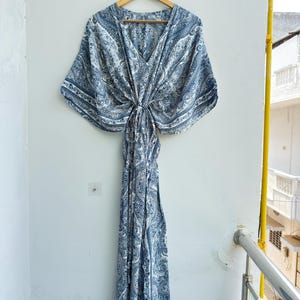 May include: A floor-length kaftan dress featuring a blue and white paisley pattern. The garment has a V-neckline, wide kimono-style sleeves, and a gathered centre waist detail that creates a draped silhouette. It is displayed on a wooden hanger.