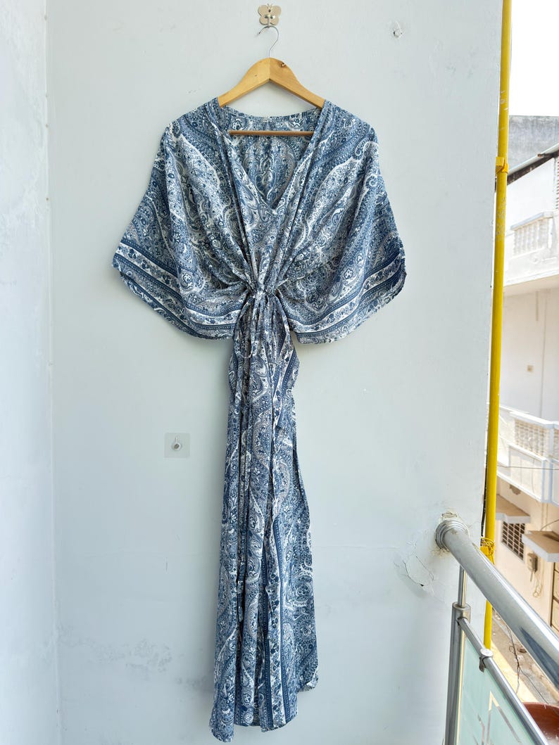 May include: A floor-length kaftan dress featuring a blue and white paisley pattern. The garment has a V-neckline, wide kimono-style sleeves, and a gathered centre waist detail that creates a draped silhouette. It is displayed on a wooden hanger.