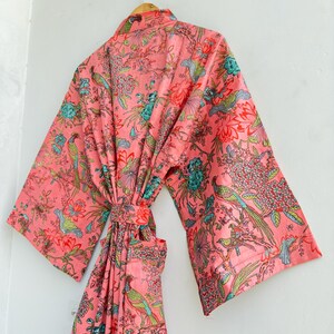 May include: A long-sleeved robe in a vibrant coral pink hue, featuring a detailed print of colourful birds, blooming flowers, and lush foliage. The garment includes a matching fabric belt at the waist and wide sleeves, hanging from a wooden hanger.