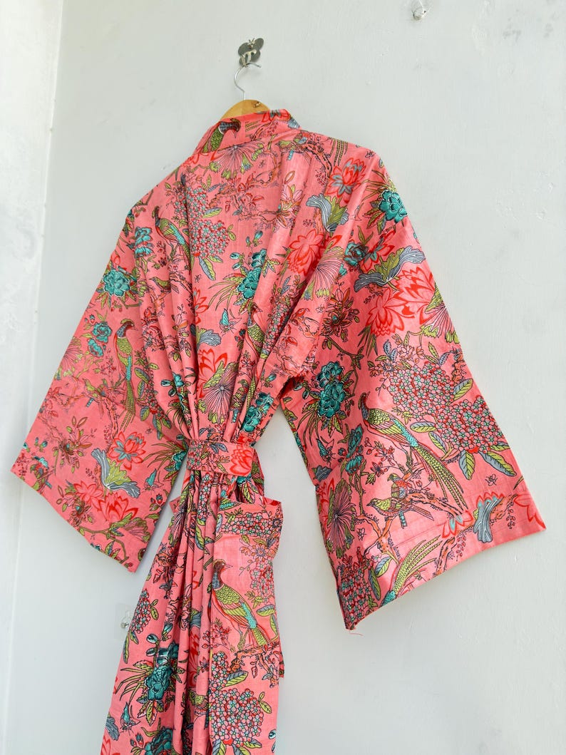 May include: A long-sleeved robe in a vibrant coral pink hue, featuring a detailed print of colourful birds, blooming flowers, and lush foliage. The garment includes a matching fabric belt at the waist and wide sleeves, hanging from a wooden hanger.
