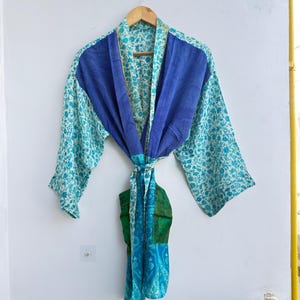 May include: A blue and teal floral print robe with a solid blue panel across the shoulders. The robe is hanging on a wooden hanger. The robe has a tie belt and is made of a lightweight fabric.
