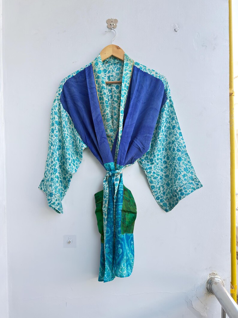May include: A blue and teal floral print robe with a solid blue panel across the shoulders. The robe is hanging on a wooden hanger. The robe has a tie belt and is made of a lightweight fabric.