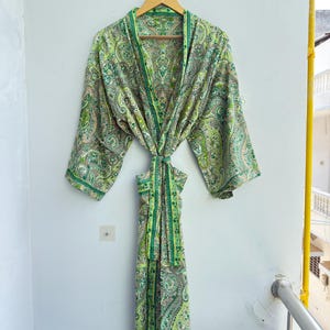 May include: A long-sleeved robe featuring a vibrant green and white paisley pattern. The garment includes a matching fabric tie belt at the waist, wide sleeves, and a shawl collar, all displayed on a wooden hanger against a plain wall.