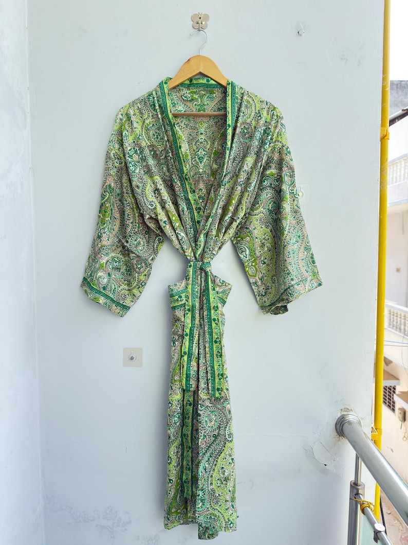 May include: A long-sleeved robe featuring a vibrant green and white paisley pattern. The garment includes a matching fabric tie belt at the waist, wide sleeves, and a shawl collar, all displayed on a wooden hanger against a plain wall.