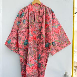 May include: A pink kimono robe featuring a repeating pattern of birds, flowers, and foliage in shades of teal, green, and red. The garment has wide sleeves, an open front with a matching fabric belt, and a relaxed, knee-length silhouette.