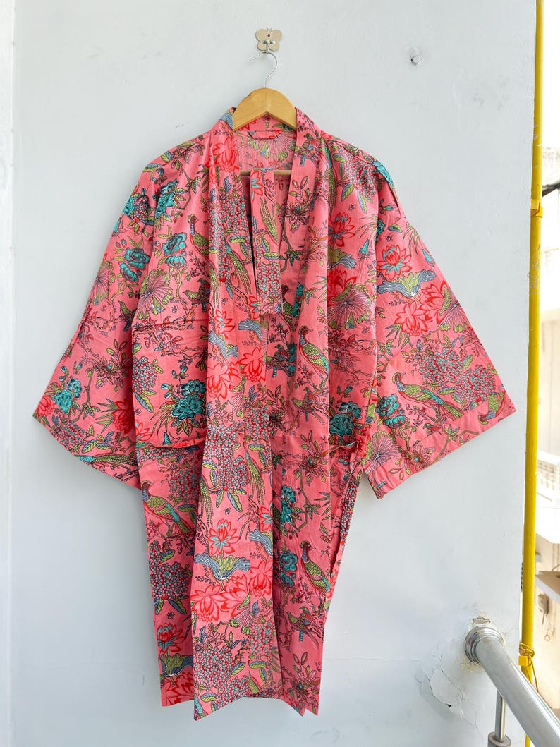 May include: A pink kimono robe featuring a repeating pattern of birds, flowers, and foliage in shades of teal, green, and red. The garment has wide sleeves, an open front with a matching fabric belt, and a relaxed, knee-length silhouette.