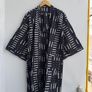 Może przedstawiać: Czarna szata w stylu kimono z białym geometrycznym wzorem kresek i kropek. Szata ma szerokie rękawy i dwie przednie kieszenie. Wisi na drewnianym wieszaku na białej ścianie.