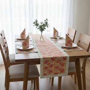 Può includere: Un tavolo da pranzo in legno apparecchiato per sei persone con un runner floreale e tovagliette abbinate. Ogni posto a tavola comprende un piatto bianco, un tovagliolo piegato e un bicchiere da vino. Il tavolo è circondato da sedie in legno.
