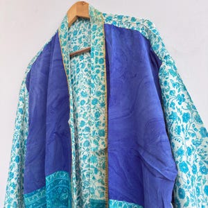 May include: A blue and white patterned kimono robe hanging on a wooden hanger. The robe features a solid blue center panel, bordered by patterned panels in shades of blue and white. The floral pattern adds a touch of elegance to the garment.