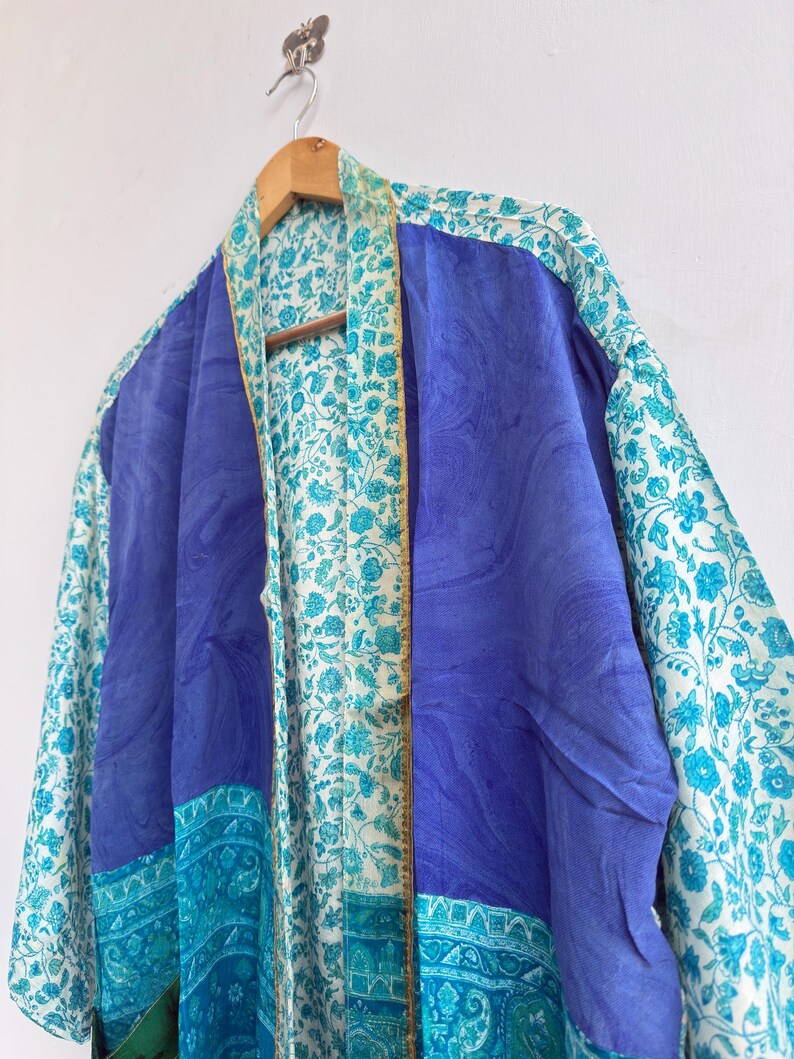 May include: A blue and white patterned kimono robe hanging on a wooden hanger. The robe features a solid blue center panel, bordered by patterned panels in shades of blue and white. The floral pattern adds a touch of elegance to the garment.