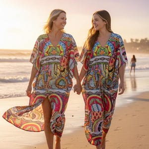 Könnte beinhalten: Zwei Frauen gehen am Strand spazieren, halten Händchen und tragen passende, farbenfrohe Maxikleider mit Muster. Die Kleider haben einen V-Ausschnitt und kurze Ärmel mit einer lebendigen Mischung aus roten, gelben, blauen und grünen Designs. Die Szene ist von warmem Sonnenlicht durchflutet.