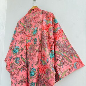 May include: A light pink kimono robe featuring a repeating pattern of vibrant teal, green, and red birds perched among detailed floral and botanical elements. The garment has wide, loose sleeves and is displayed on a wooden hanger.