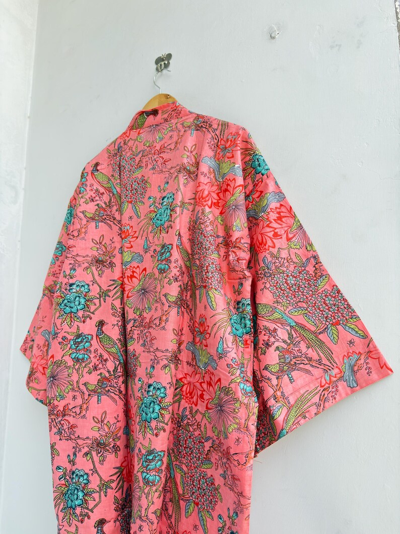 May include: A light pink kimono robe featuring a repeating pattern of vibrant teal, green, and red birds perched among detailed floral and botanical elements. The garment has wide, loose sleeves and is displayed on a wooden hanger.