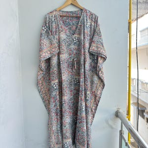 Könnte beinhalten: Ein langer, fließender Kaftan mit V-Ausschnitt und Kordelzug in der Taille. Der Kaftan hat ein detailliertes, allover Muster in Grau-, Rot- und Cremetönen. Er hängt an einem Holzbügel vor einer weißen Wand und zeigt sein lockeres Design.