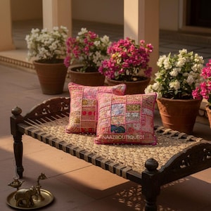 Könnte beinhalten: Zwei rosafarbene Patchwork-Kissen liegen auf einer dunklen Holzbank mit gewebter Sitzfläche. Die Kissen haben eine Vielzahl von Stoffquadraten in Rosa- und Rottönen. Die Bank steht vor Topfpflanzen mit rosa und weißen Blüten.
