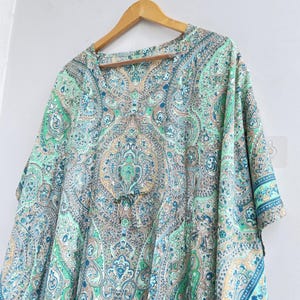 May include: A loose-fitting tunic featuring a vibrant paisley pattern in shades of mint green, teal, brown, and cream. The garment has a V-neckline, short sleeves, and a central drawstring detail at the waist, all displayed on a wooden hanger.