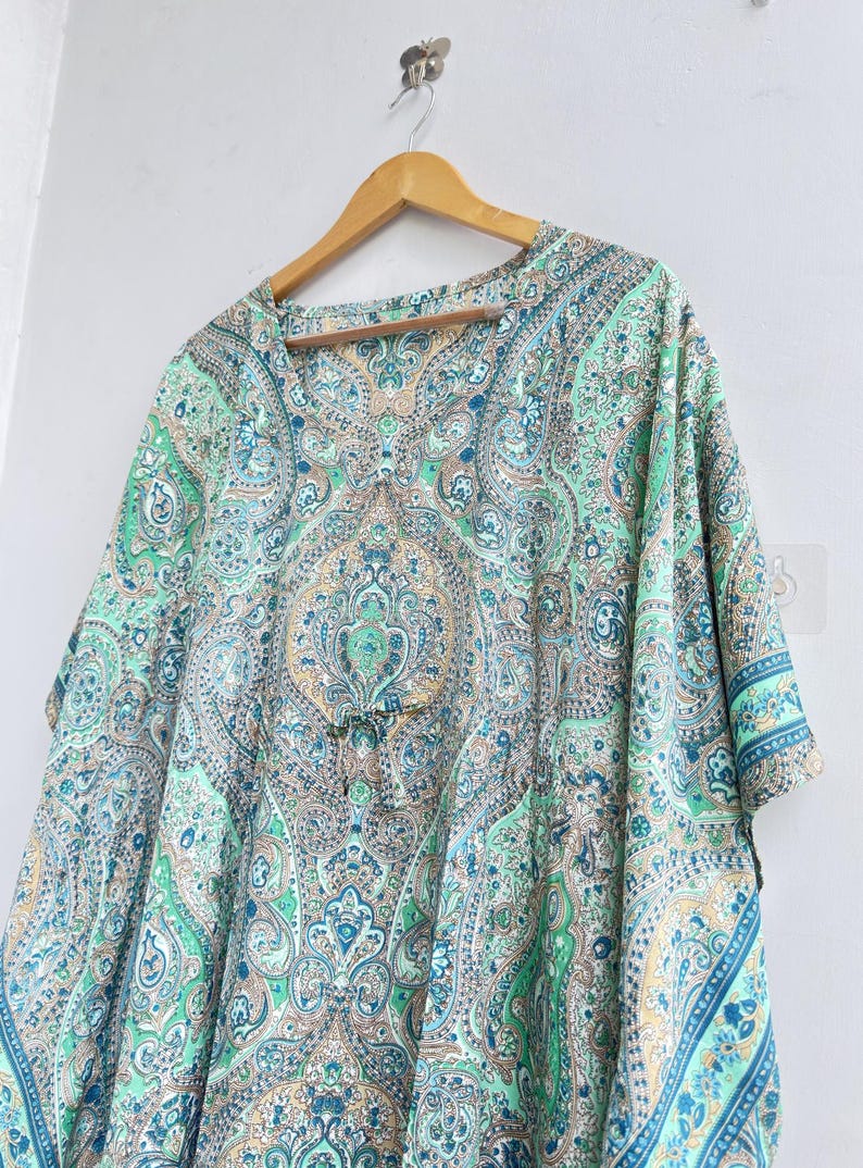 May include: A loose-fitting tunic featuring a vibrant paisley pattern in shades of mint green, teal, brown, and cream. The garment has a V-neckline, short sleeves, and a central drawstring detail at the waist, all displayed on a wooden hanger.