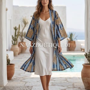 Könnte beinhalten: Eine Frau trägt ein langes, fließendes weißes Kleid und einen blau-gold gemusterten Kimono. Der Kimono hat ein Tigerstreifen-Design. Das Bild wurde im Freien aufgenommen, mit einem Pool und Pflanzen im Hintergrund. Der Text "NAAZBOUTIQUE01" ist sichtbar.