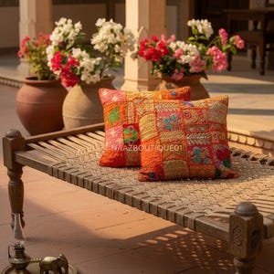 Könnte beinhalten: Zwei quadratische orangefarbene Zierkissen mit bunten Patchwork-Designs und gestickten Details liegen auf einem gewebten Bett. Die Kissen sind im Freien in der Nähe von Topfblumen platziert. Der Text "NAAZBOUTIQUE01" ist auf einem Kissen zu sehen.
