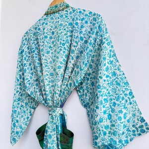 May include: A light blue and white floral print kimono robe hanging on a wooden hanger. The robe is tied at the waist with a matching sash. The robe has long sleeves and a green and blue patterned trim.