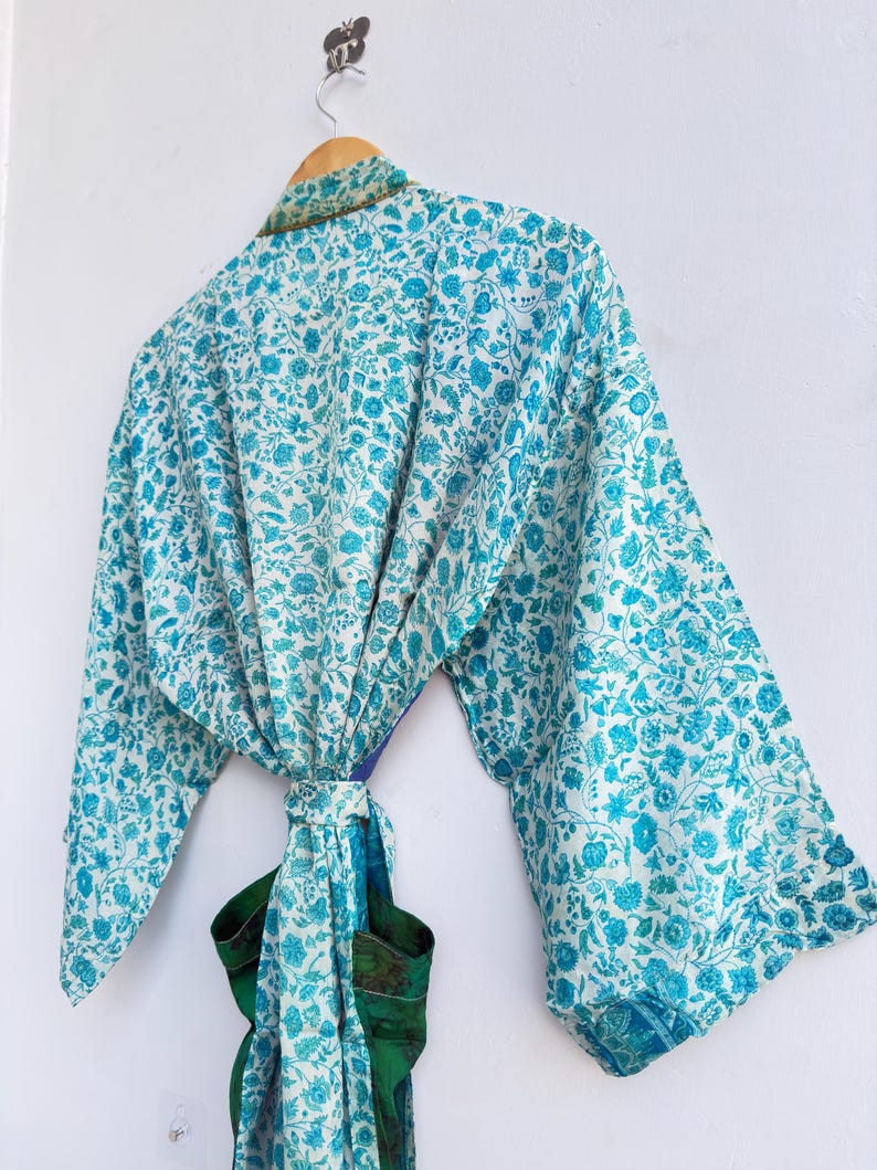 May include: A light blue and white floral print kimono robe hanging on a wooden hanger. The robe is tied at the waist with a matching sash. The robe has long sleeves and a green and blue patterned trim.