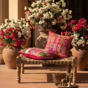 Op de afbeelding: Twee decoratieve roze patchwork kussens liggen op een kleine houten bank. De bank staat voor grote terracotta potten gevuld met witte en rode bloeiende planten. Een kleine messing lade met een olifantenbeeldje staat op de grond.