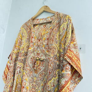 May include: A loose-fitting kaftan featuring a vibrant paisley pattern in shades of yellow, orange, and white. The garment has a V-neckline, short sleeves, and a drawstring detail at the centre front, all displayed on a wooden hanger.