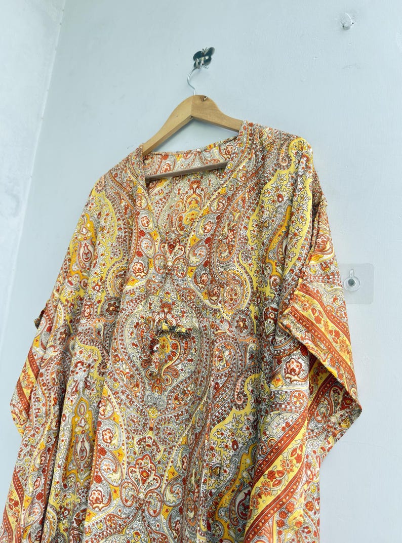May include: A loose-fitting kaftan featuring a vibrant paisley pattern in shades of yellow, orange, and white. The garment has a V-neckline, short sleeves, and a drawstring detail at the centre front, all displayed on a wooden hanger.