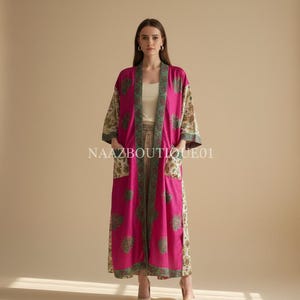May include: A long, flowing magenta kimono with floral patterned sleeves and pocket details. The kimono has a green and cream floral trim and the text "NAAZBOUTIQUE01" is visible. The model is wearing a cream top and matching pants.