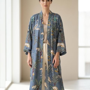 May include: A long, open-front blue kimono with a jungle print featuring leopards, trees, and birds. The kimono is worn over a cream-colored top and white pants. The text "NAAZBOUTIQUE01" is visible at the bottom.
