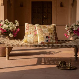 Könnte beinhalten: Eine Holzbank mit gewebter Sitzfläche trägt zwei dekorative quadratische Kissen. Die Kissen haben ein Patchwork-Design in Gold-, Beige- und Rottönen. Bougainvillea-Blüten in Rosa und Weiß befinden sich in großen Terrakotta-Töpfen.