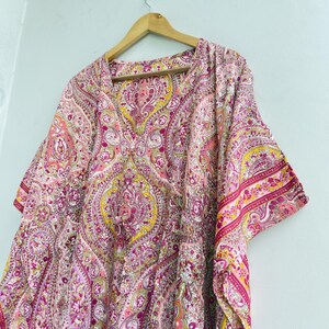May include: A kaftan featuring a vibrant paisley pattern in shades of pink, magenta, yellow, and white. The garment has a V-neckline, wide short sleeves, and a drawstring waist detail, hanging from a wooden hanger against a plain light background.