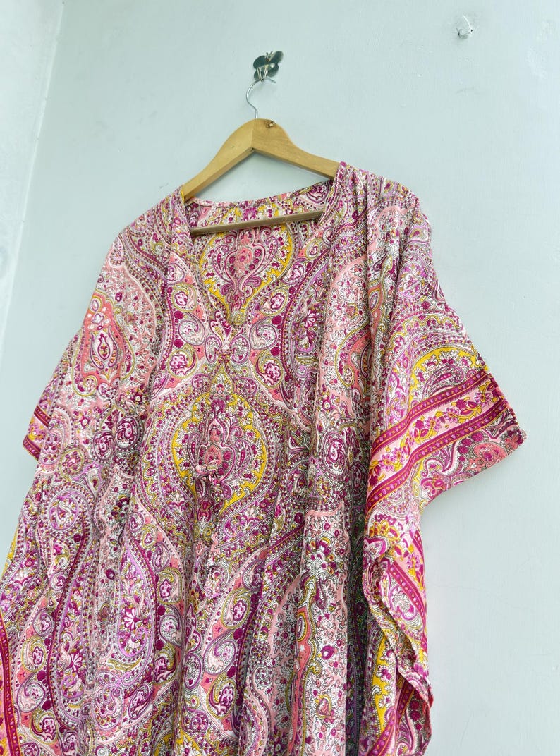 May include: A kaftan featuring a vibrant paisley pattern in shades of pink, magenta, yellow, and white. The garment has a V-neckline, wide short sleeves, and a drawstring waist detail, hanging from a wooden hanger against a plain light background.