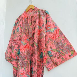 May include: A long-sleeved kimono-style robe in a vibrant pink hue, featuring an all-over print of colourful birds, blooming flowers, and lush foliage in shades of teal, green, and red. The garment has a wide collar and a matching fabric belt.