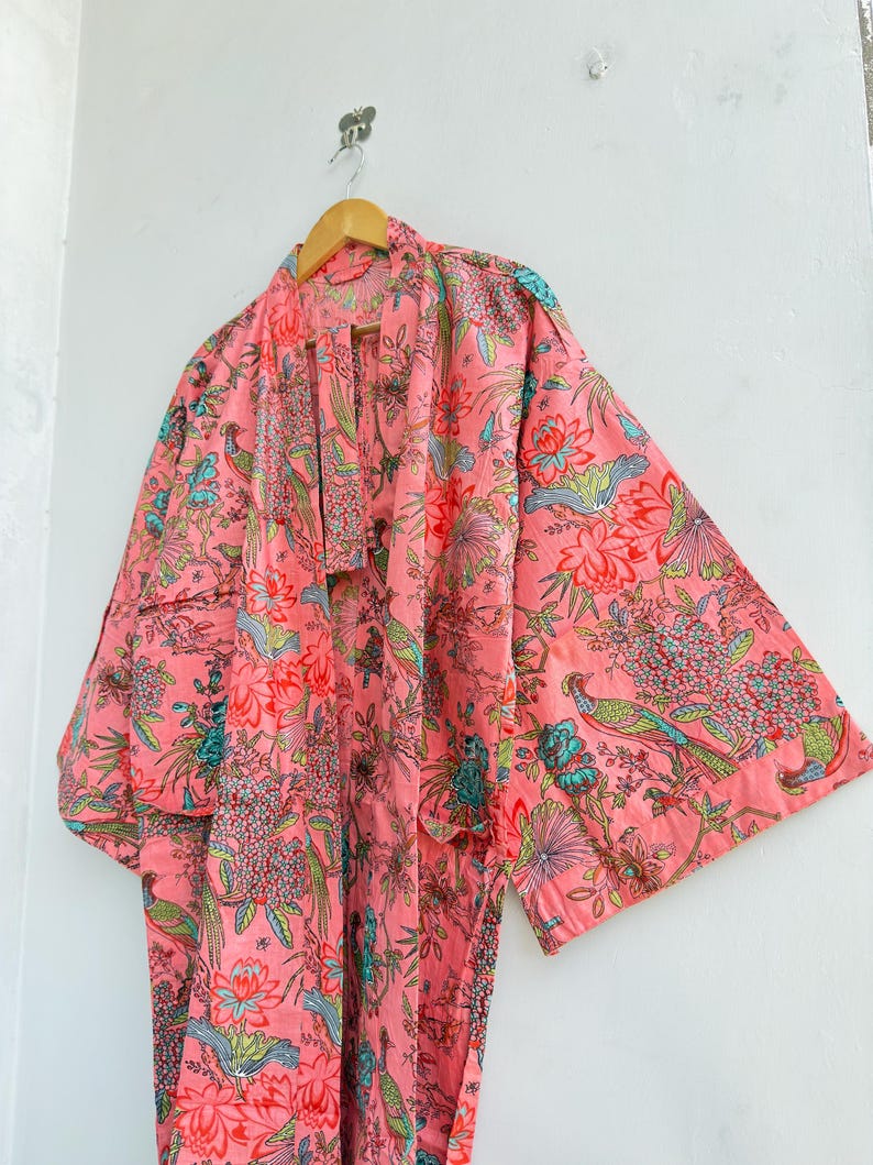 May include: A long-sleeved kimono-style robe in a vibrant pink hue, featuring an all-over print of colourful birds, blooming flowers, and lush foliage in shades of teal, green, and red. The garment has a wide collar and a matching fabric belt.