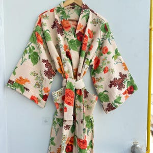 Könnte beinhalten: Ein cremefarbener Blumen-Bademantel mit Bindegürtel. Der Bademantel hat ein Muster aus roten, orangefarbenen und grünen Blumen und Blättern. Der Bademantel hängt an einem Holzbügel vor einer weißen Wand.