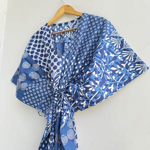 Könnte beinhalten: Ein blau-weiß gemustertes Kaftankleid hängt an einem Holzkleiderbügel. Das Kleid weist verschiedene geometrische und florale Designs in Blau- und Weißtönen auf. Der Kaftan ist mit einem passenden Stoffgürtel in der Taille gebunden.