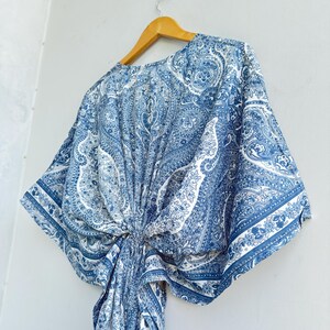 May include: A blue and white paisley print kaftan top featuring wide kimono-style sleeves and a gathered knot detail at the centre front. The garment is displayed on a wooden hanger, showcasing a repeating intricate pattern throughout the fabric.