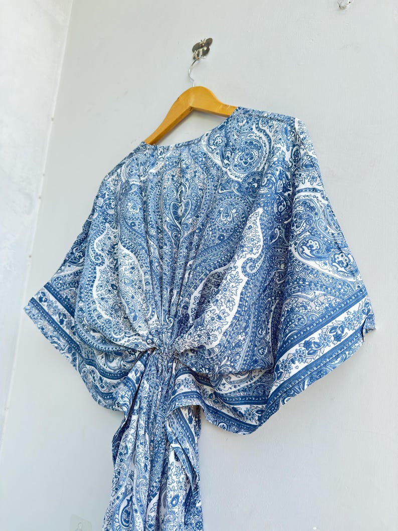 May include: A blue and white paisley print kaftan top featuring wide kimono-style sleeves and a gathered knot detail at the centre front. The garment is displayed on a wooden hanger, showcasing a repeating intricate pattern throughout the fabric.