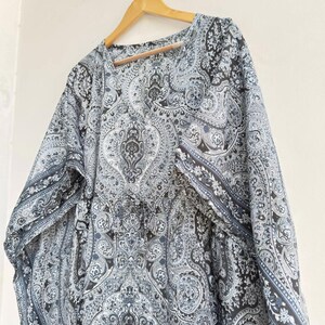 May include: A loose-fitting kaftan featuring a detailed black, white, and grey paisley pattern throughout. The garment has a V-neckline, wide sleeves, and a drawstring waist detail with small tassels, all displayed on a wooden hanger.