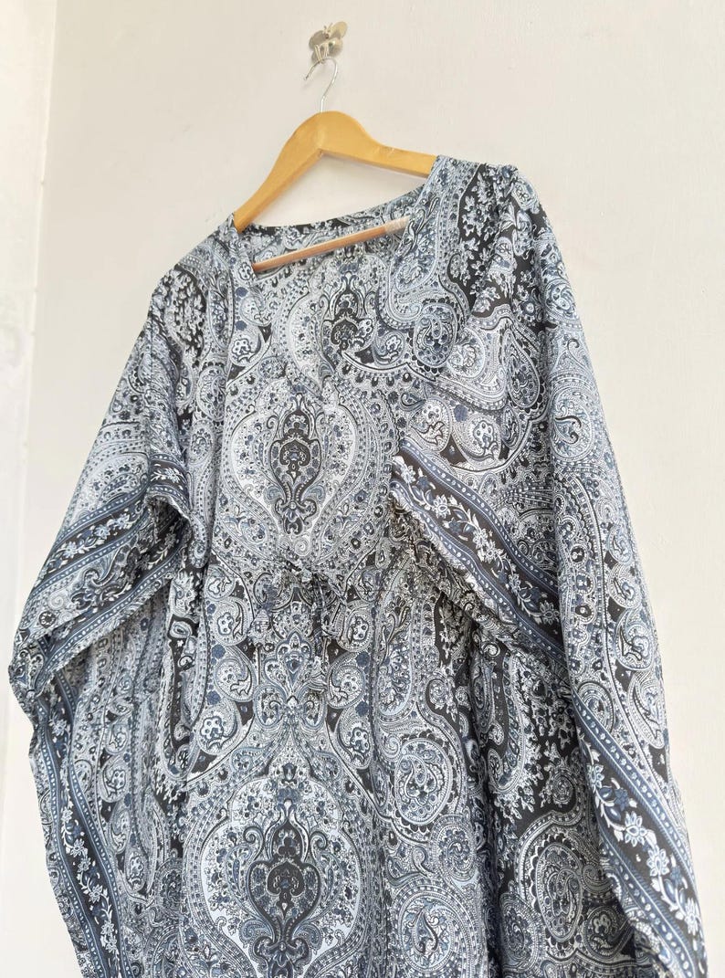 May include: A loose-fitting kaftan featuring a detailed black, white, and grey paisley pattern throughout. The garment has a V-neckline, wide sleeves, and a drawstring waist detail with small tassels, all displayed on a wooden hanger.