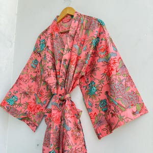 May include: A coral pink robe featuring a vibrant floral and bird pattern in shades of teal, green, and white. The garment has wide, loose sleeves, a wrap-front design with a matching fabric tie belt, and a shawl collar, hanging on a wooden hanger.