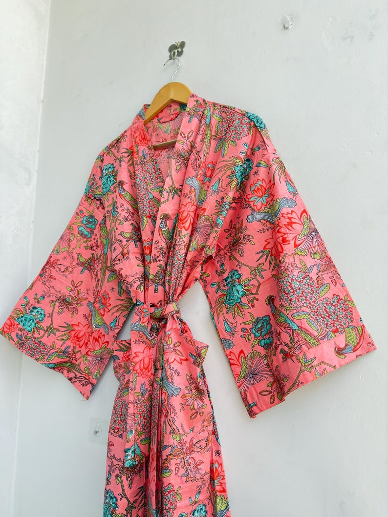 May include: A coral pink robe featuring a vibrant floral and bird pattern in shades of teal, green, and white. The garment has wide, loose sleeves, a wrap-front design with a matching fabric tie belt, and a shawl collar, hanging on a wooden hanger.