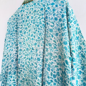May include: A floral patterned kimono in shades of white and turquoise hangs on a wooden hanger. The kimono features a detailed design of flowers and leaves. The collar is trimmed with a gold-colored fabric.