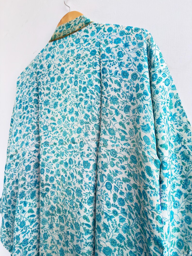 May include: A floral patterned kimono in shades of white and turquoise hangs on a wooden hanger. The kimono features a detailed design of flowers and leaves. The collar is trimmed with a gold-colored fabric.