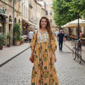 Könnte beinhalten: Ein langes, senfgelbes Kleid mit vertikalen weißen Streifen und grünen Blumenmustern. Das Kleid hat einen V-Ausschnitt mit floraler Stickerei und Puffärmeln. Die Person trägt braune Sandalen und eine braune Handtasche. Der Hintergrund ist eine Kopfsteinpflasterstraße.
