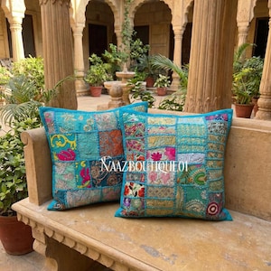 Puede incluir: Dos cojines cuadrados de patchwork con borde turquesa. Los cojines presentan una variedad de cuadrados de tela coloridos con estampados florales y geométricos. El texto "VAAZBOUTIQUE01" es visible en un cojín.