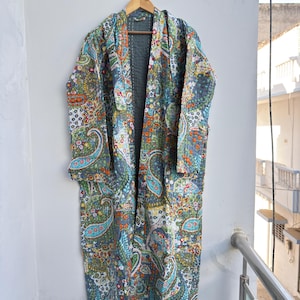 Könnte beinhalten: Ein langer, offen geschnittener Bademantel mit einem farbenfrohen Paisley-Muster in Grau-, Blau-, Orange- und Grüntönen. Der Bademantel hängt an einem Holzbügel vor einer weißen Wand. Der Bademantel scheint aus einem gesteppten Stoff zu sein.