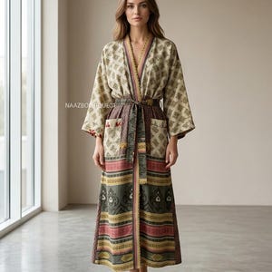 Könnte beinhalten: Langer, gemusterter Bademantel mit V-Ausschnitt und passendem Gürtel. Der Bademantel hat einen beigefarbenen Hintergrund mit einem sich wiederholenden geometrischen Muster, und der untere Teil hat eine Mischung aus roten, grünen und braunen Streifen und Designs. Der Text "NAAZBOUTIQUE01" ist sichtbar.